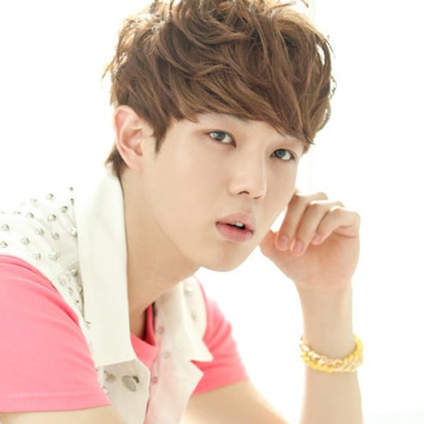 Iri has brown hair. He is wearing a white vest that's adorned with fake rhinestones, and a light pink shirt underneath. He also wears a curly yellow bracelet coupled with a clear one.
