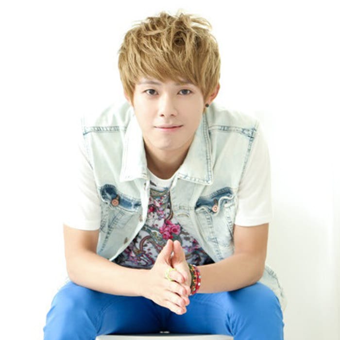 Woosu has golden blond hair. He is wearing a light blue denim vest with a white, floral-patterned t-shirt underneath. He also wears 3 bracelets: one red, one pink, and one green. They all appear to have colourful rhinestones on them. On his right index finger, he dons a neon yellow ring. He wears neon blue pants as well.