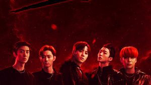he male members of the group SHAX alongside a red and black background. They wear dark clothing. They are standing in a line facing front. From left to right, its Leehyun, Jaewoo, KwonRyuk, Dojin and finally Hyuk. Above the members, there is the group logo 'SHAX' in a fancy black and red design.