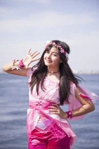 Mutsumi waving at the camera in front of the ocean. she is wearing a pink outfit.