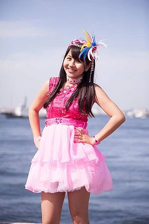 yukaren stood in front of the ocean. she has both hands on her hips, and is wearing a pink outfit.