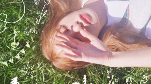 aylah laying on grass, covering her eyes from the sun with her hand. there is graphic text of the word 'anomaly' in the bottom left corner